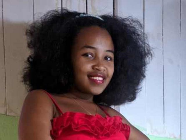 SandSexy with dark brown skin, curly black hair, red ruffled dress, smiling, showing small teeth, wearing a thin necklace, sitting against a white wooden wall.