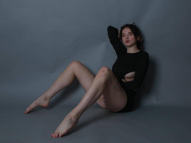 EllaFerrera with pale skin, short dark hair, black long-sleeve shirt, no pants, sitting on gray floor, legs spread, arm behind head, tattoos on thigh, bare feet, relaxed.