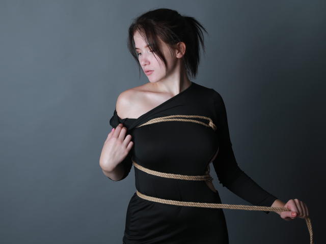 EllaFerrera with rope binding her tits and arms, black off-shoulder dress, dark brown hair in a messy ponytail, holding rope, grey background, slight smile, looking to the side.