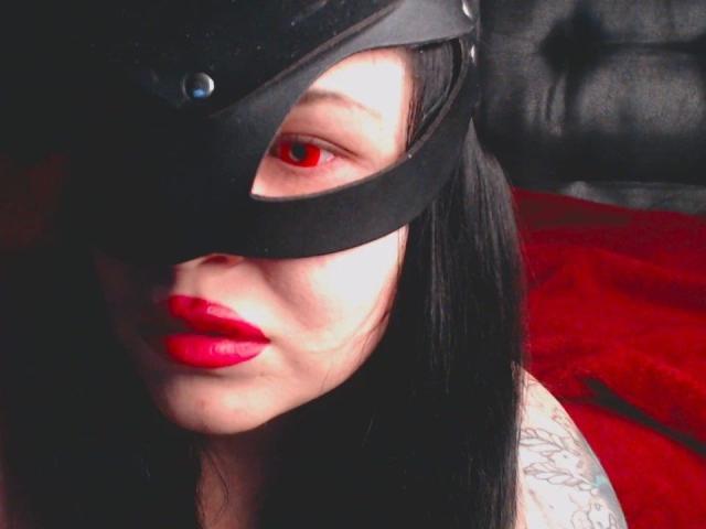 Psychobitch7: una donna attraente con una maschera nera che copre un occhio, rossetto rosso, spalla tatuata, capelli scuri e pelle chiara, in un contesto rosso e nero, che guarda seducentemente la telecamera..