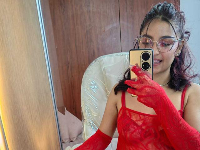 Ameliebx with glasses, red lace dress, and red mesh gloves, holding phone, smiling, showing small tits, and medium brown skin, reflected in mirror.