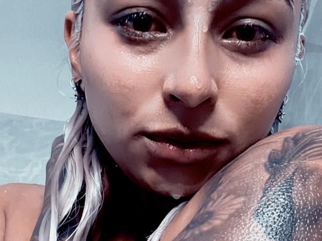 AvrilGray has wet, blonde braids, a tattooed shoulder, and a glistening face with cum on her nose and chin. She's looking up with a slightly open mouth.