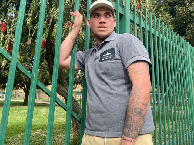LuckyStar69 with tattoos on a tattooed arm, wearing a gray polo, beige cap, and beige pants, leaning on a green metal fence, looking slightly bored.