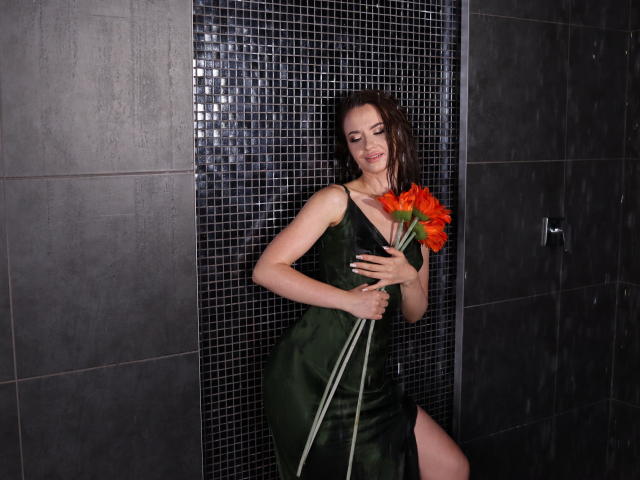 KaraCriegs in a green satin dress holding orange flowers against her tits, smiling with wet brown hair, standing in a dark tiled shower.