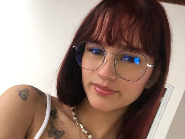 MelodyMansson with red hair, glasses, and a white top, showing tattoos on her shoulder and chest, smiling with pink lips, and wearing a pearl necklace.