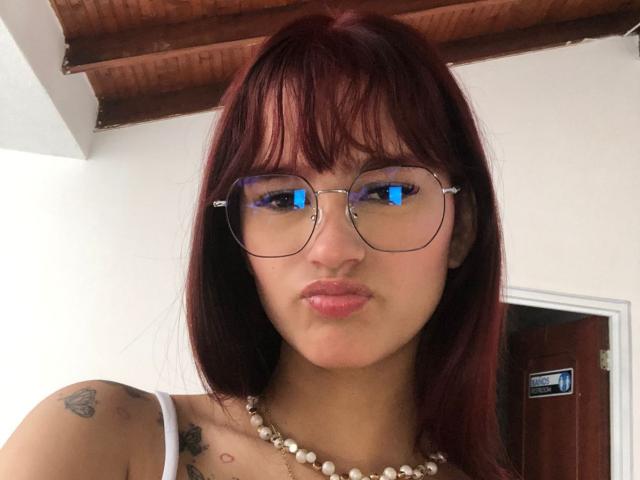 MelodyMansson with red hair, glasses, pink lips, white tank top, pearl necklace, tattoos, and reflection in glasses, standing in a room with wooden ceiling.