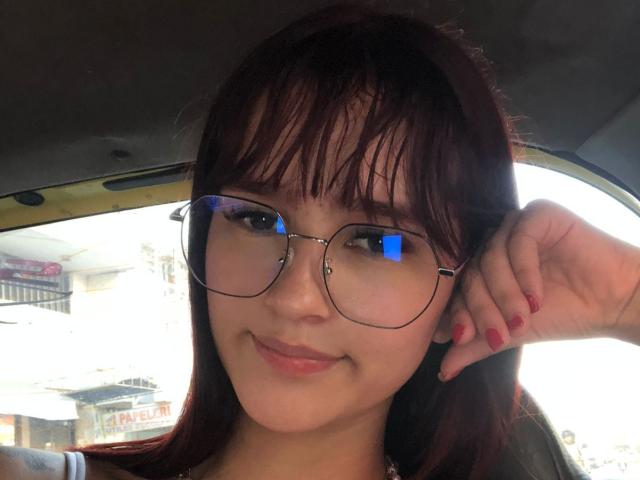 MelodyMansson with glasses, red nail polish, and dark brown hair, smiling in a car, her right hand near her ear. Reflections on her glasses. Soft light on her face.