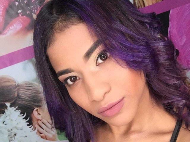 Zafiira with purple hair, medium brown skin, and subtle makeup, looking directly at the camera with a slight smile. Her tits are not visible. Her lips are slightly parted.
