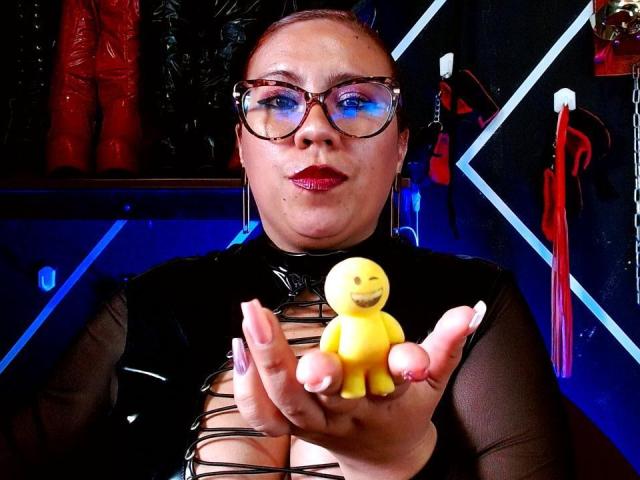 LexyDom with glasses, red lipstick, and black lace-up top, holding a yellow plastic baby doll in her hand. She's got a chubby face, and her nails are long and pink.