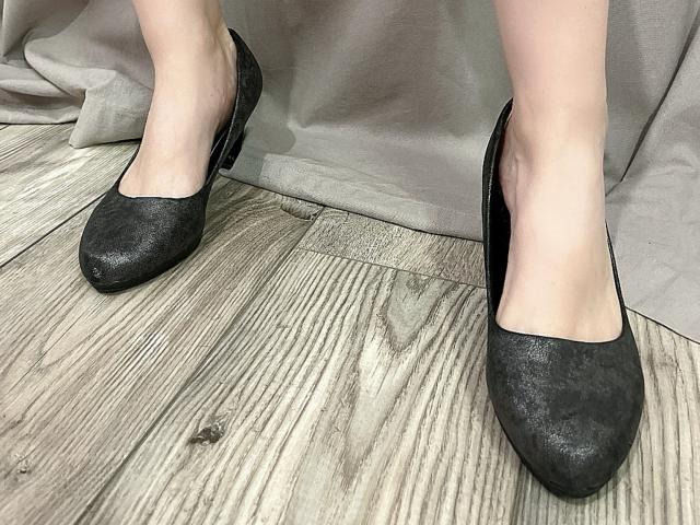 MelanieFrancaise's feet in black, textured flats with a slightly worn look, standing on a wooden floor with visible grain. Her pale skin contrasts with the dark shoes.