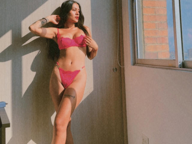 ThaliaAres stands in pink lace lingerie, her hands touching her tits, with long dark hair, tattoos, and a sexy, confident look, in sunlight near a window.