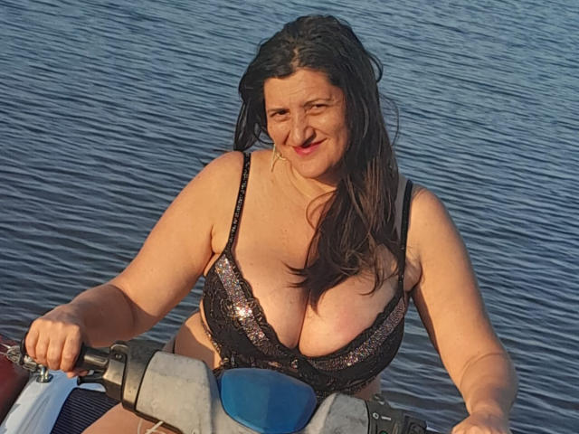 BlissSophia with wet black sequined bikini top, showing big boobs, smiling on a boat, holding the handle, blue water in background, dark brown hair, fair skin.