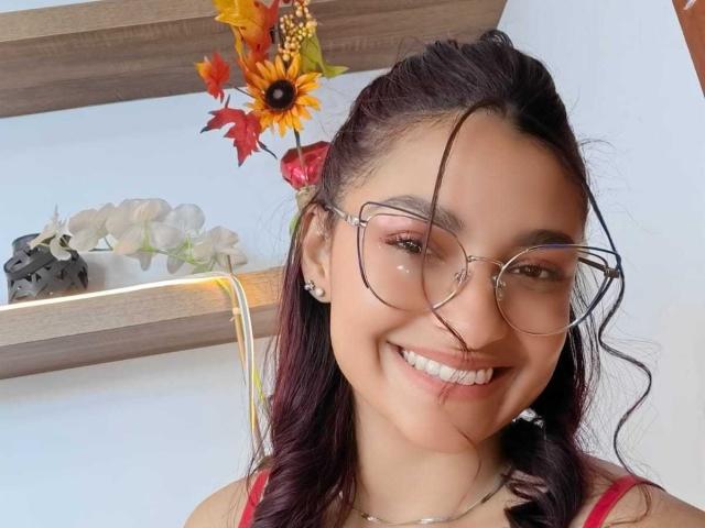 Ameliebx, avec des lunettes, souriante, cheveux noirs ondulés, haut rouge, dents blanches et boucles d'oreilles, tenant un tournesol et des feuilles d'automne. Ses seins sont petits, et elle est mignonne..