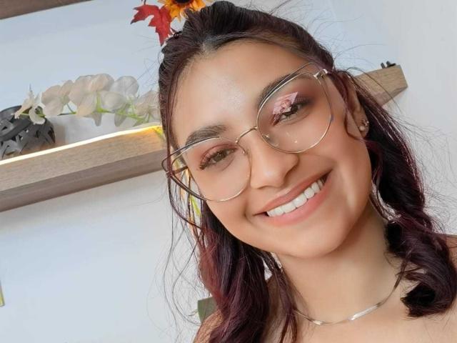 Ameliebx, portant des lunettes, souriante, montrant des dents blanches, avec un collier argenté, des cheveux roux foncés et un fard à paupières rose discret. Les seins ne sont pas visibles, l'accent est mis sur le visage..