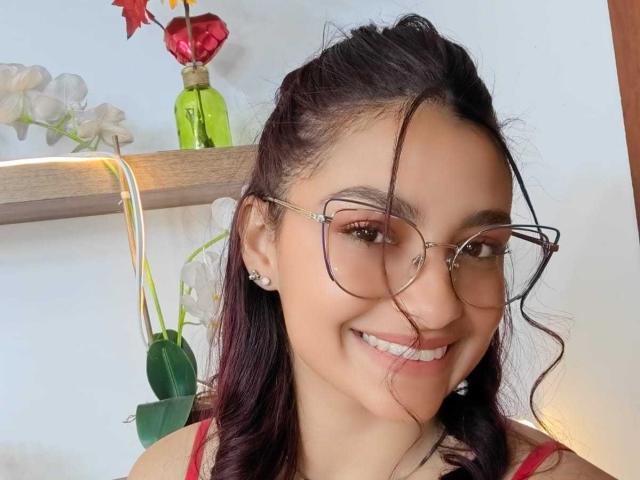 Ameliebx with dark wavy hair, glasses, nose ring, earrings, smiling, showing teeth, wearing red top, white wall, green vase, red flower, white orchids in background.