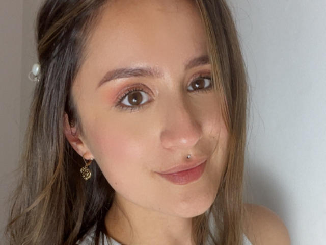 NinaBryons has brown eyes, light brown hair, and a small nose piercing. She's wearing gold earrings with a floral design and a subtle smile. Her skin is fair.