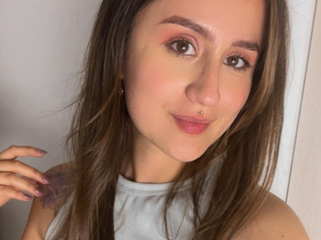 NinaBryons has brown eyes, pink eyeshadow, a nose piercing, and a slight smile. She's wearing a white sleeveless top, with long brown hair and a tattoo on her left arm.