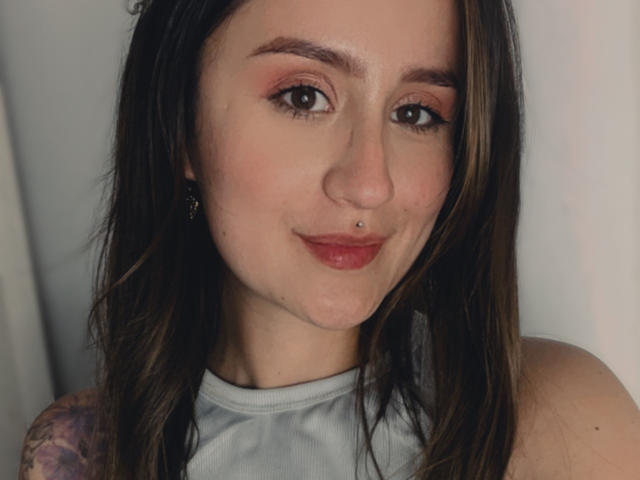 NinaBryons has light skin, brown eyes, pink eyeshadow, red lipstick, a nose piercing, and a tattoo on her left shoulder. She's smiling with her boobs barely visible.