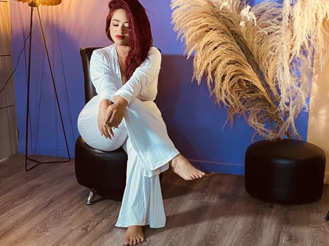 HannaMiillys with red hair, white dress, and bare feet, sitting on black chair, legs crossed, puffy pants, relaxed, soft smile, pampas grass, warm light, wood floor.