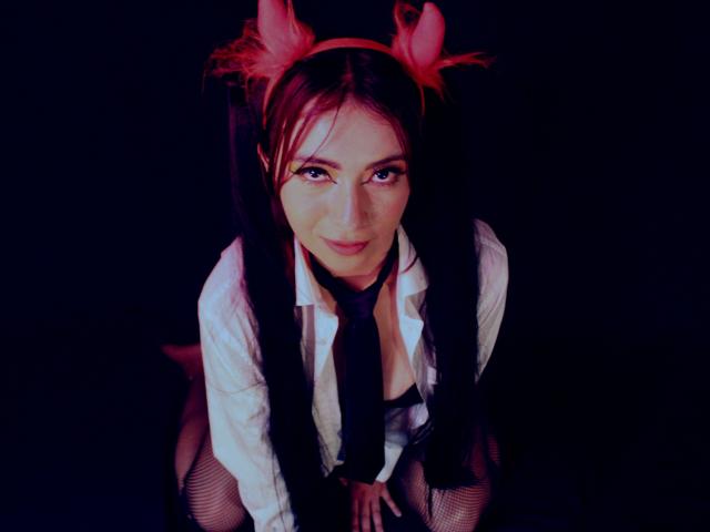 AnaGerald, avec des oreilles de lapin rouges, des couettes foncées, une chemise blanche, une cravate noire et des bas en résille, regardant vers le haut avec un sourire joueur, les mains sur les genoux, dans un décor sombre..