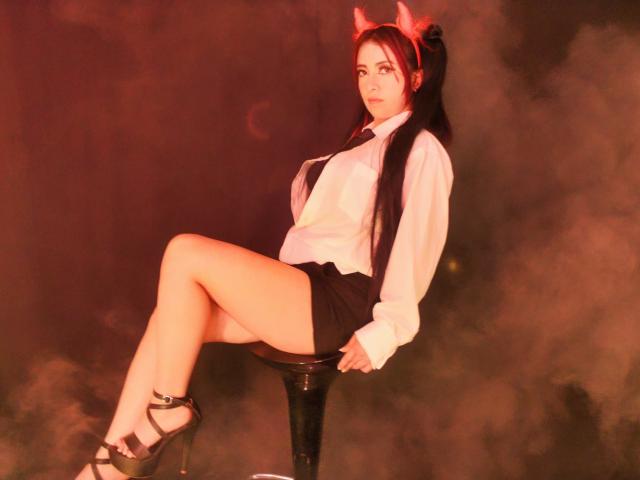 AnaGerald mit roten Teufelshörnern, weißem Hemd, schwarzen Shorts, High Heels, sitzend auf einem Hocker, Beine gekreuzt, langes, dunkles Haar, ernster Gesichtsausdruck, rotes Licht, sichtiges Gesäß, sexy..