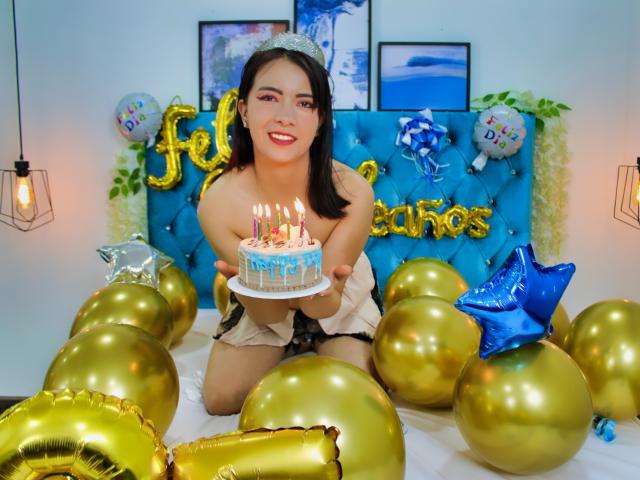 AnaGerald, con i capelli neri, sorridente, che tiene una torta con le candeline, senza indumenti superiori, seduta su un letto circondata da palloncini dorati e blu. I suoi piccoli seni sono visibili..