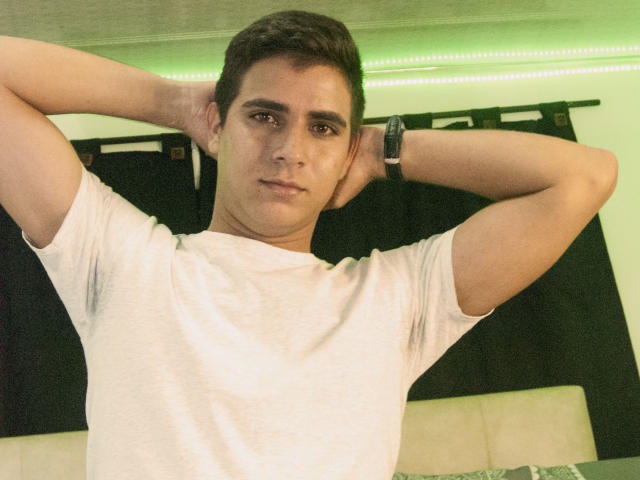 SantosMontenegro with hands behind head, wearing white tee, wristwatch, short brown hair, green-lit background, slightly serious expression, medium skin, cute, casual.