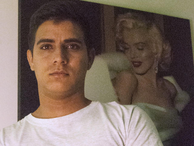 SantosMontenegro has short dark hair, brown eyes, and a serious expression. He's wearing a white shirt. Behind him is a picture of Marilyn Monroe's face and shoulders.