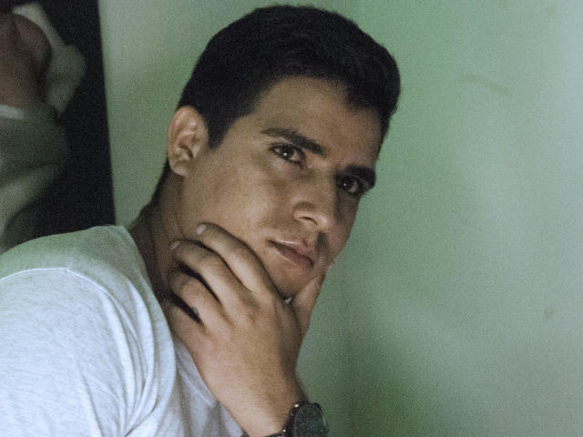 SantosMontenegro, with short dark hair and brown eyes, resting his chin on his hand, wearing a white shirt, looking slightly to the side with a serious expression.