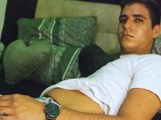 SantosMontenegro is lying on a bed, wearing a white shirt, with a green and patterned pillow behind him. His wrist has a black watch, and he's looking at the camera.