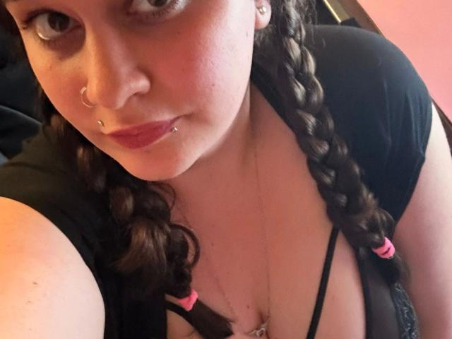 SweetGirl1998-hot with braided brown hair, black shirt, and pink hair ties, nose ring, and lip piercing, showing a slight smile, and her large, exposed tits in the photo.