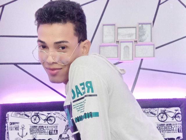 NickSull with short curly hair, wearing clear glasses and a gray hoodie with teal text, smiling over shoulder. His ass is not visible.