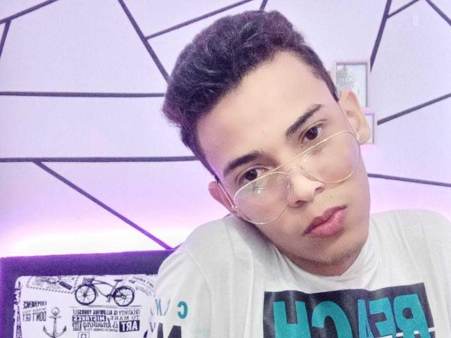 NickSull with short dark hair, wearing clear glasses, white shirt with green text, pouty lips, and a relaxed expression, in a purple-lit room.