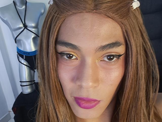 NickSull's face with long brown hair, gold glittery eyeliner, brown eyes, pink lipstick, and a silver mannequin torso in the background. Boobs not visible, focus on NickSull's face.