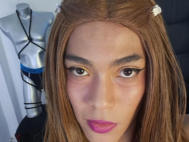 NickSull has light brown skin, straight brown hair with white clips, thick eyebrows, glittery brown eyes, and pink lipstick. Silver mannequin with black straps is in the background.