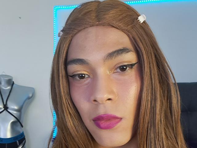 NickSull tiene el cabello castaño, cejas pobladas y usa lápiz labial rosa. Sus párpados tienen brillo y delineador de ojos negro. A la izquierda, hay un maniquí con correas negras..