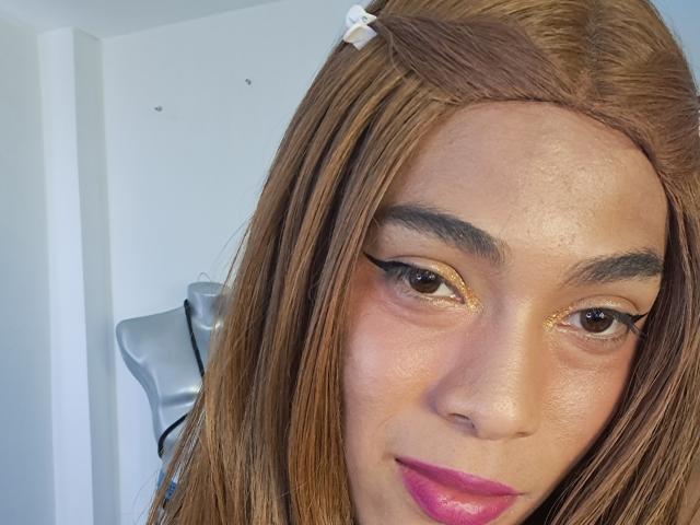 NickSull has long brown hair, gold eyeshadow, black eyeliner, pink lipstick, and a white hairpin. Her skin is light brown with visible pores. Her brown eyes look at the camera.