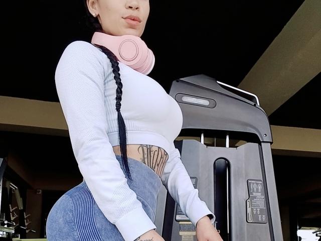 EvaMueller with pink headphones, white long-sleeve crop top, blue high-waisted jeans, visible tattoo on side, standing by a gray treadmill, looking down, slightly pouty face.