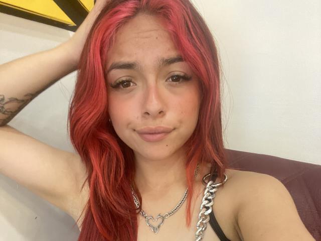 Yovglau, con i capelli rossi, gli occhi marroni e un piccolo sorriso, indossa una collana a catena e ha un braccio alzato, mostrando un tatuaggio sul braccio sinistro. Il seno è visibile..