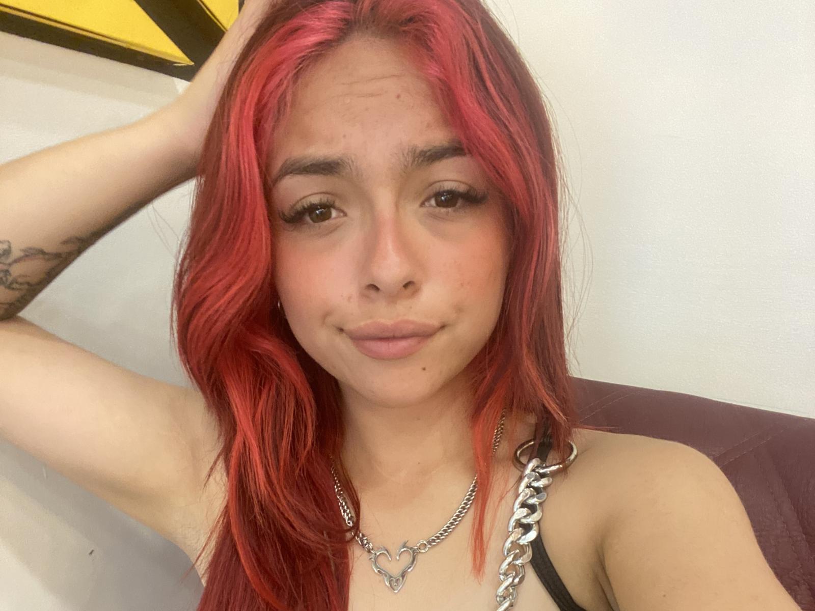 Yovglau with red hair, brown eyes, and a small smile, wearing a chain necklace, has her arm raised, showing a tattoo on her left arm. Boobs visible.