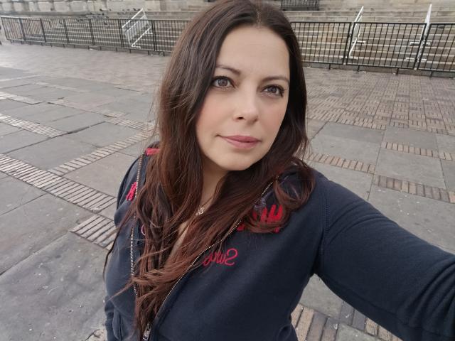 Seher tem cabelos castanhos longos, pele clara e olhos castanhos. Ela está vestindo um moletom preto com zíper e bordados vermelhos. Seus seios são de tamanho médio. Ela está tirando uma selfie ao ar livre..
