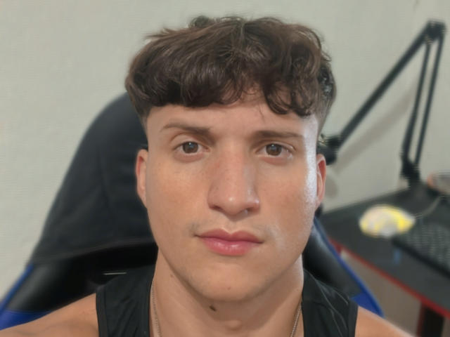 TheRiqque has a cute face with brown wavy hair, brown eyes, and pink lips. He's wearing a black sleeveless shirt and a gold chain, sitting in a chair.