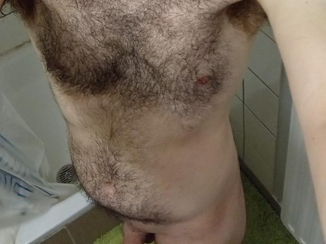 El pecho peludo de Aure69, con pelo oscuro y grueso en las axilas y el pecho, pequeños pezones rosados y un abdomen peludo que sobresale ligeramente, está de pie en un baño con una alfombra verde..