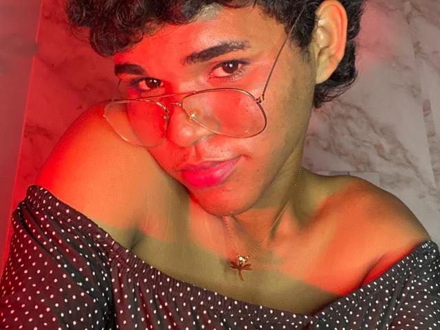 AlexaaCute with curly hair, wearing glasses, pink lipstick, and a black off-shoulder polka dot top, necklace with a star pendant, red light on face.