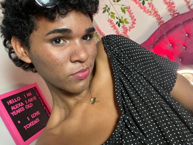AlexaaCute with curly black hair, brown skin, pink lipstick, black polka dot shirt, necklace, and a slight smile, looking at the camera. Visible pink headboard and green leaves.