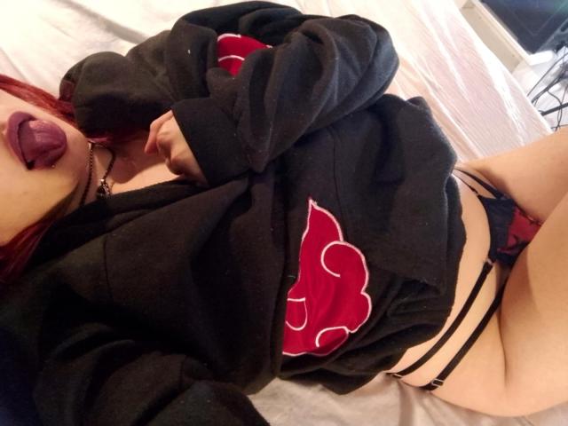 MaiaThunder liegt auf einem Bett, die Zunge herausgestreckt, trägt eine schwarze Kapuzenjacke mit einem roten und weißen Logo, schwarze Slips mit dünnen Trägern und eine schwarze Halskette, hat rotes Haar und blasse Haut..