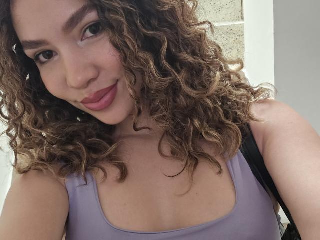 KendallPiierce69 with curly brown hair, light skin, wearing a purple tank top, showing a slight smile, and looking at the camera, with a black strap over her shoulder.