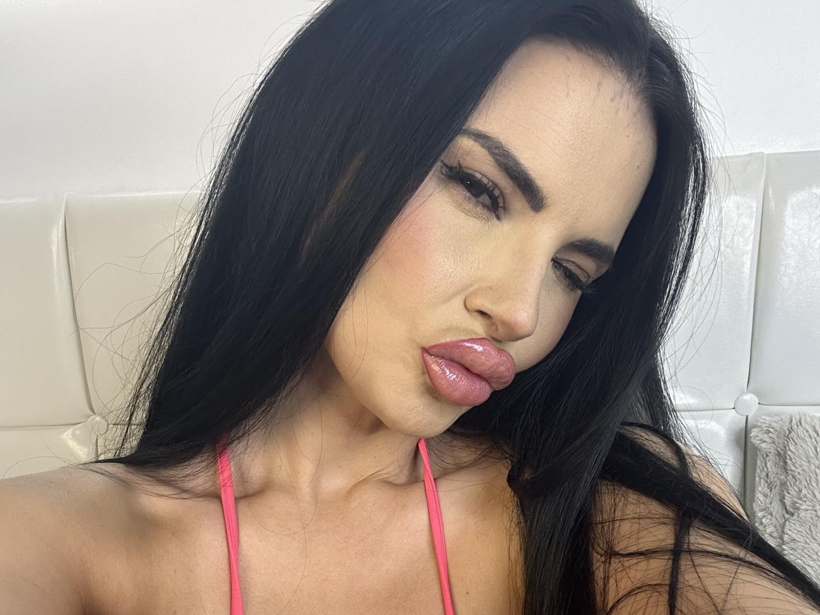 NicolleHeaven mit langen schwarzen Haaren, pinkem Lippenstift und herausgesteckter Zunge, trägt ein pinkes Halstopp und zeigt ihre Brüste. Sie hat dunkle Augenbrauen und Eyeliner..