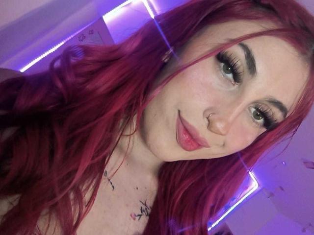 SammyViolet has red hair, a nose ring, and tattoos on her chest, with puffy red lips and long eyelashes, looking at the camera with a slight smile.
