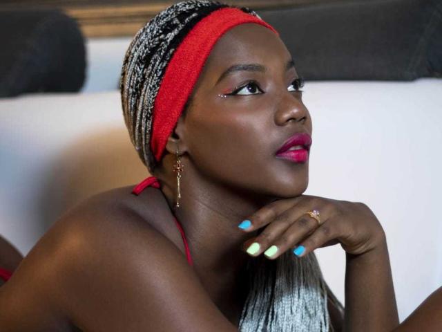 KaiaJhons with dark skin, red headband, black-and-white pattern, bright pink lipstick, blue nail polish, gold earrings, ring, and her hand resting on her chin, looking to the side.